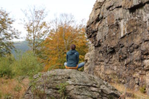 Eine Frau sitzt im Schneidersitz auf einem Felsen vor einem herbstlich gefärbtem Baum