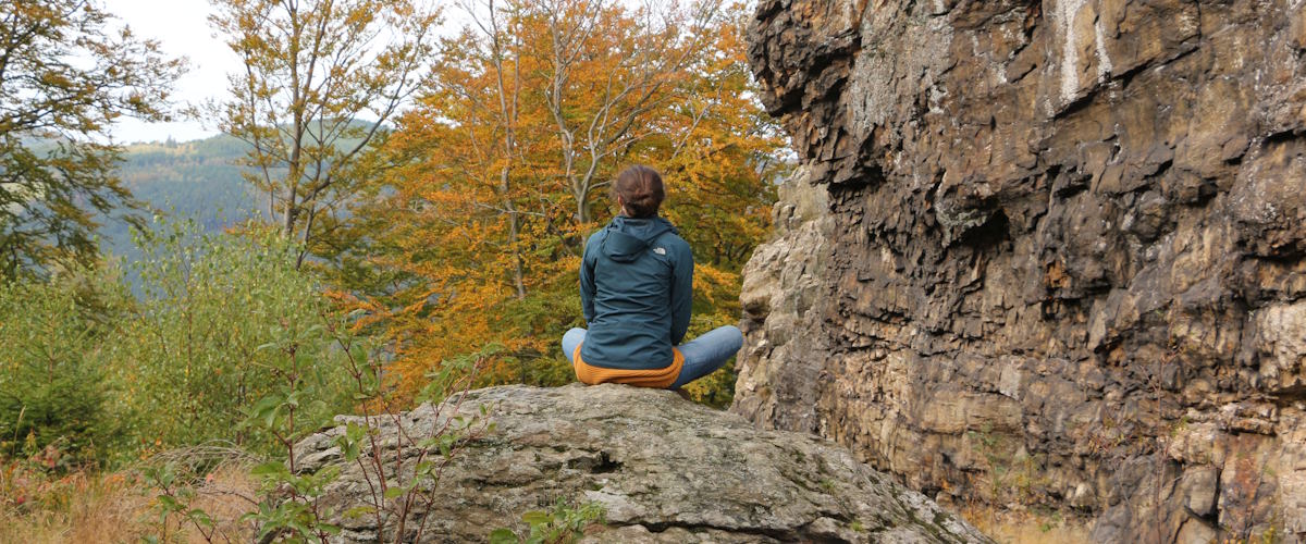 Eine junge Frau sitzt im Schneidersitz auf einem Stein, im Hintergrund ein Herbstbaum mit orange gefärbtem Laub