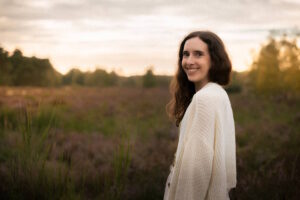 Ayurveda Ernährungsberaterin und Gesundheitsberaterin Miriam steht in einer blühenden Heide, sie trägt die Haare offen und einen weißen Strickcardigan
