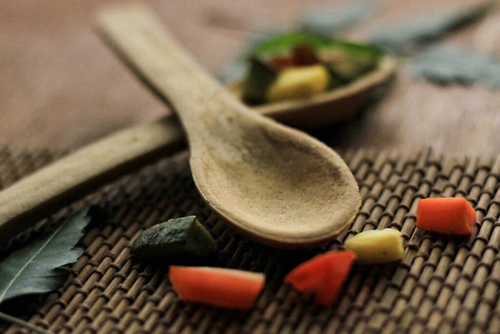 Auf einem Tisch liegen zwei Holzlöffel und ayurvedische Kräuter und Lebensmittel wie ein Stück Ingwer, was gut ist bei einem schwachen Verdauungsfeuer Agni
